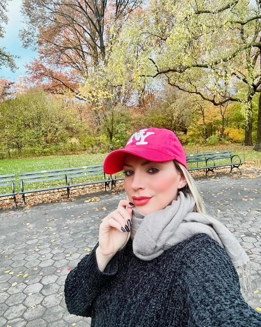 Duda Veiga em Nova York, com boné rosa, cachecol cinza e árvores ao fundo